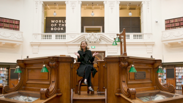 A smiling woman in a black dress sits in a timber dais in a large, elegant room