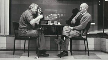 A black and white image of two men sitting outside a coffee shop sipping drinks and smoking