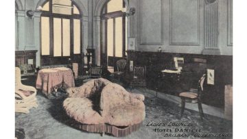 Postcard image of a ladies' lounge in the Hotel Daniell, Brisbane. The lounge has a fireplace and includes some pink armchairs and a dining table draped in a pink tablecloth