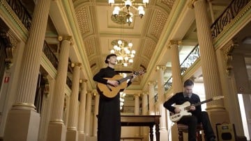 A woman and man play guitar in a grand, two-storeyed Victorian room