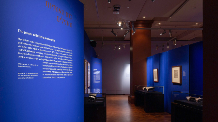 Installation view of Luminous: A thousand years of Hebrew manuscripts at State Library Victoria. View of a gallery section with cobalt blue walls and text in white and gold about the power of letters and words. There are three glass cases with rare books and manuscripts down a corridor and framed works on the walls.