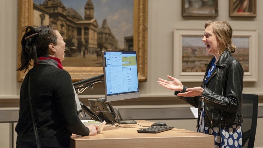 Two women laugh at a service desk in an art gallery