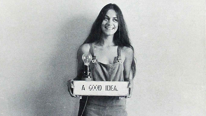 A long-haired, smiling woman in overalls holds up a sign saying 'A good idea'