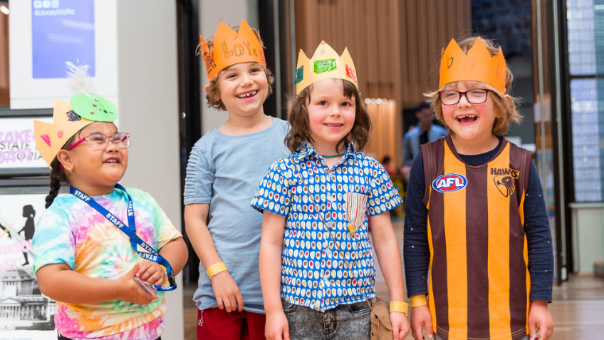Four children laughing and smiling while wearing paper crowns in the Library