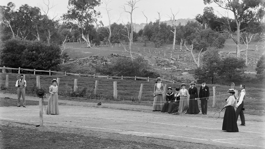 Men and women in straw hats, trousers and long skirts play tennis in bush surrounds