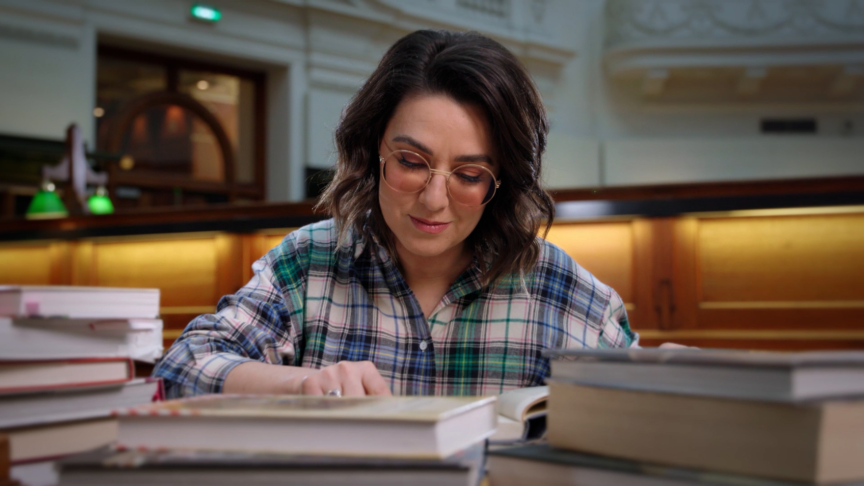 Alice Zaslavsky surrounded by stacks of books while seated a desk in the Library's La Trobe Reading Room