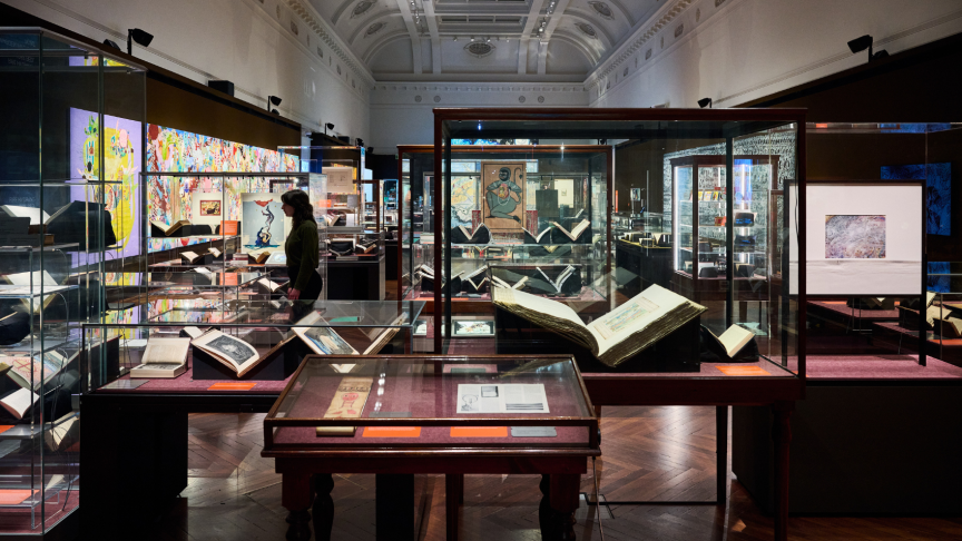 Installation view of Creative Acts: Artists and their inspiration at State Library Victoria. There are a number of cabinets and cases in the foreground featuring books and other items from the collection. It is a very full gallery.
