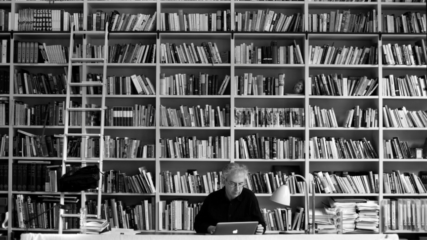 Black and white photograph of author Peter Carey working on a laptop in his study, which feature floor-to-ceiling bookshelves spanning one wall.
