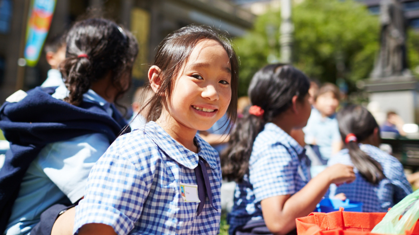 Photograph of a primary school student looking at the camera. She is on a school excursion at State Library Victoria, and other students from her school are in the background. 