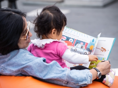 A baby being held by her mother whilst holding a picture book and turning the pages.