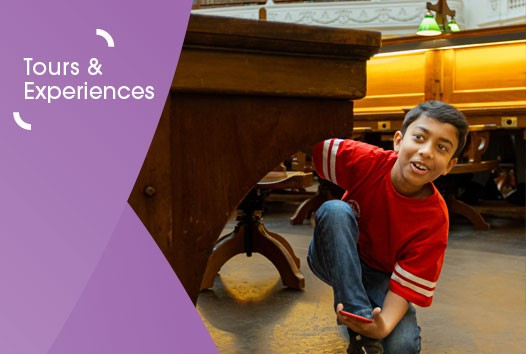 Image of a boy in a red t-shirt and blue jeans crouched under a table in the La Trobe Reading Room while holding a phone in his hand. On the left of the image is a purple graphic overlay with white text that reads 'Tours and Experiences'.