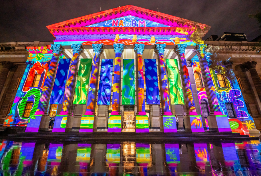A grand building with large, Ancient Greek-style pillars. Projected onto the building are colourful graphics