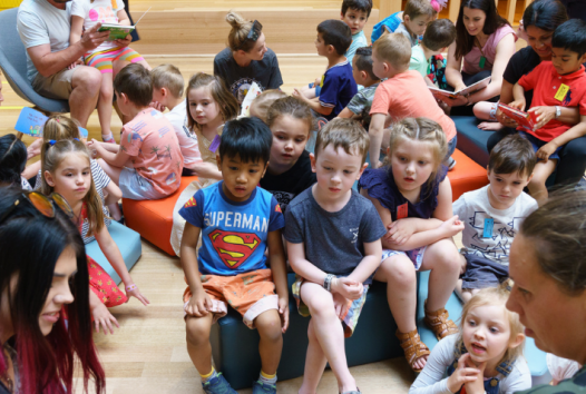 A group of young children sitting on the floor in the Library listening to a story be read out loud.
