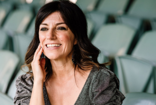 Award-winning journalist and documentary filmmaker Angela Pippos, smiling looking off camera whilst sitting in a stadium